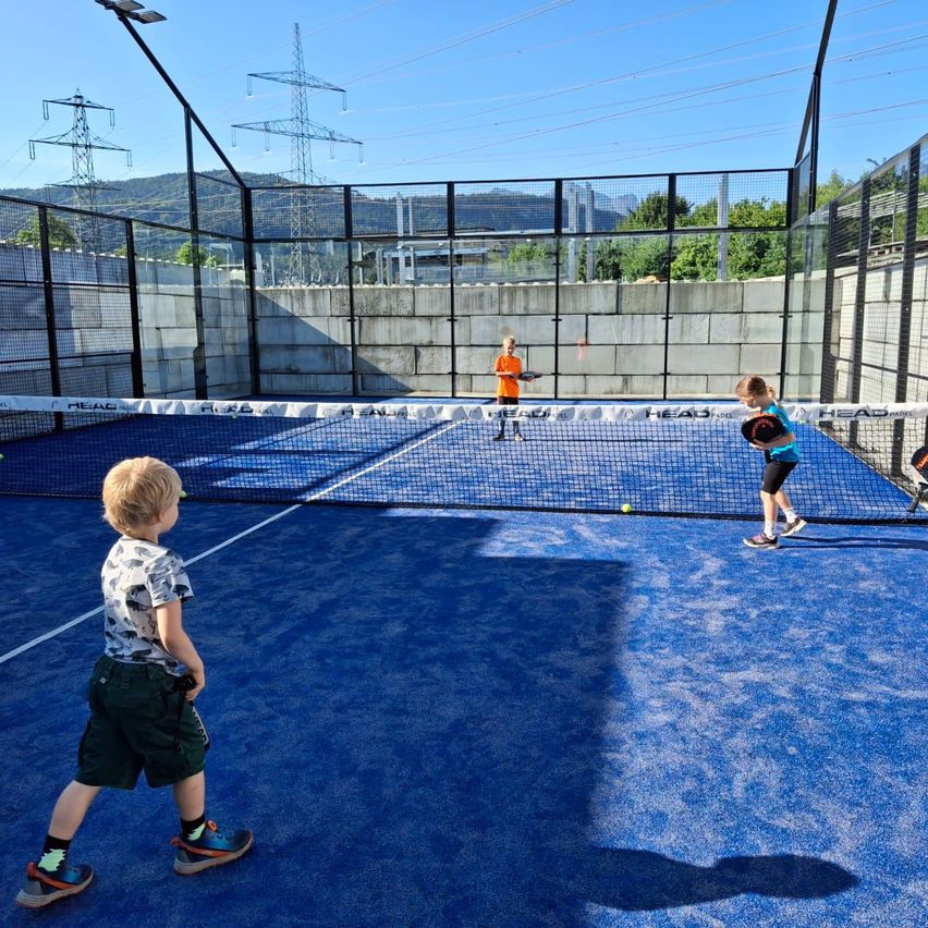 Drei Kinder spielen Tennis auf einem blauen Platz, der von einem Zaun umgeben ist, wobei ein Kind zuschaut und zwei spielen.