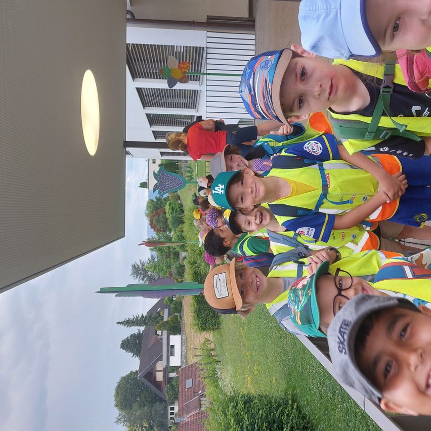Eine Gruppe von Kindern in Sicherheitswesten steht draußen, mit mehreren Erwachsenen und Bäumen im Hintergrund.