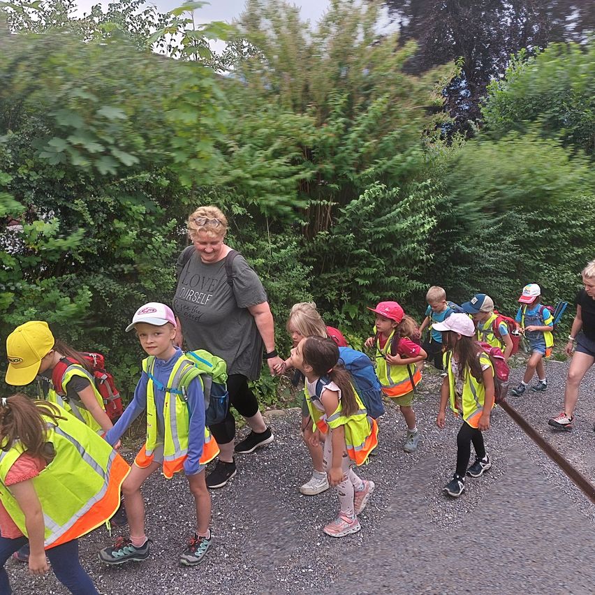 Eine Gruppe von Kindern in Sicherheitswesten geht auf einem Kiesweg. Eine Frau, wahrscheinlich ihre Lehrerin, geht neben ihnen. Bäume und Büsche befinden sich an der Seite.