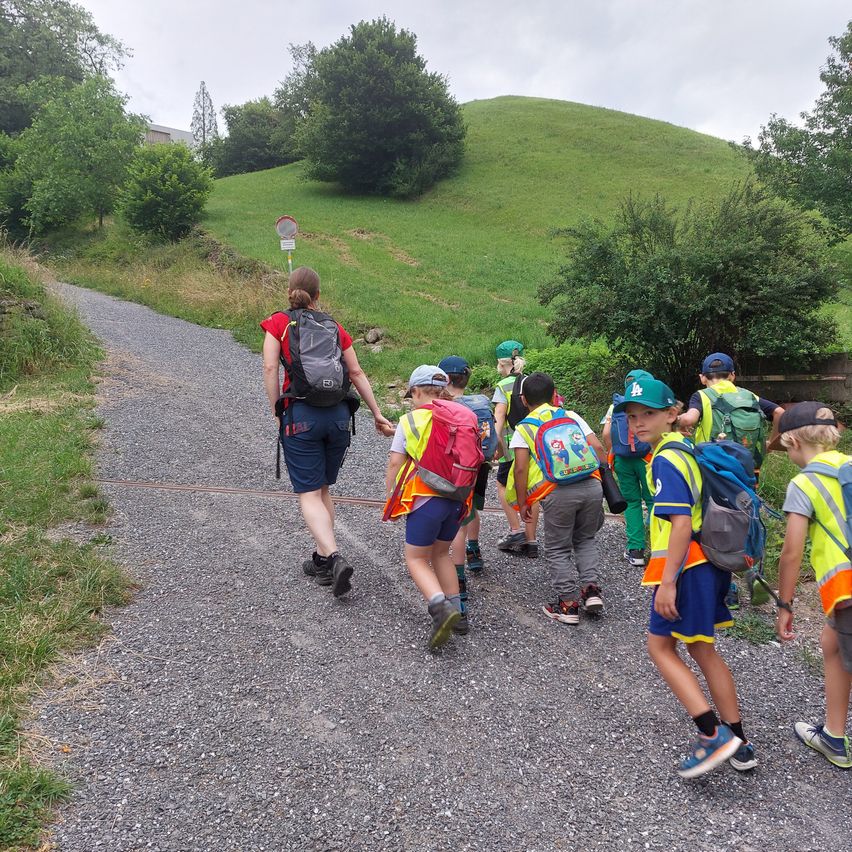 Eine Gruppe von Kindern mit reflektierenden Westen und Rucksäcken geht auf einem Kiesweg, mit einer Frau vorne. Grüne Bäume und Büsche säumen den Weg, mit einem Grashügel in der Ferne.