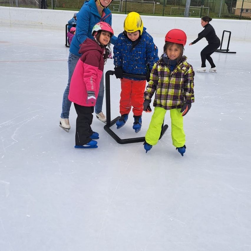 Mehrere Kinder üben das Eislaufen auf einer Eisbahn. Eine Frau hilft zwei Kindern, und ein Junge fährt im Hintergrund.