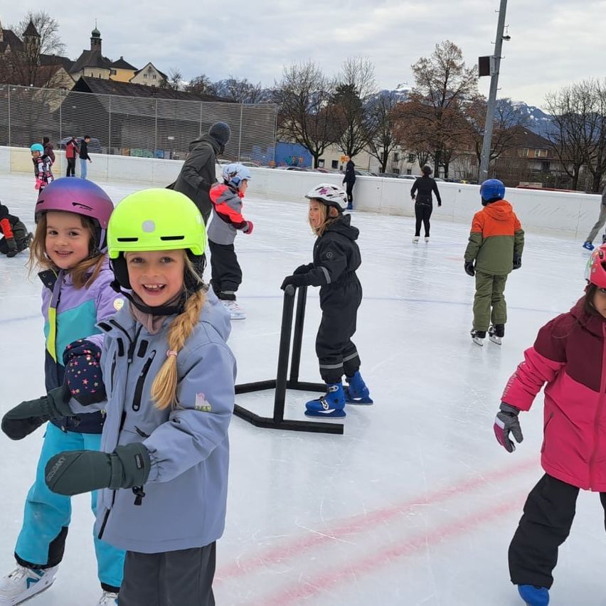 Kinder mit Helmen auf einer Eisbahn im Freien, umgeben von anderen und einer weißen Barriere. Bäume und Gebäude im Hintergrund.