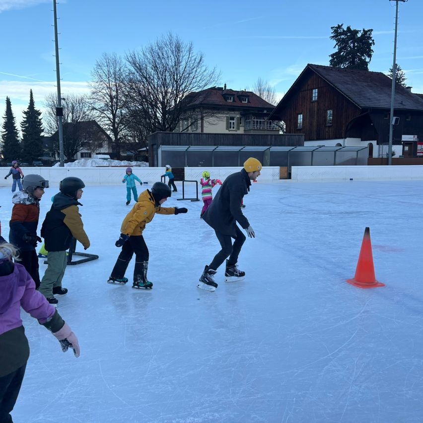 Kinder und Erwachsene fahren Schlittschuh auf einer Freiluftbahn, wobei ein Mann mit gelber Mütze und eine Frau in einem lila Mantel zusehen. Im Hintergrund befinden sich mehrere Gebäude, darunter ein großes braunes Haus.