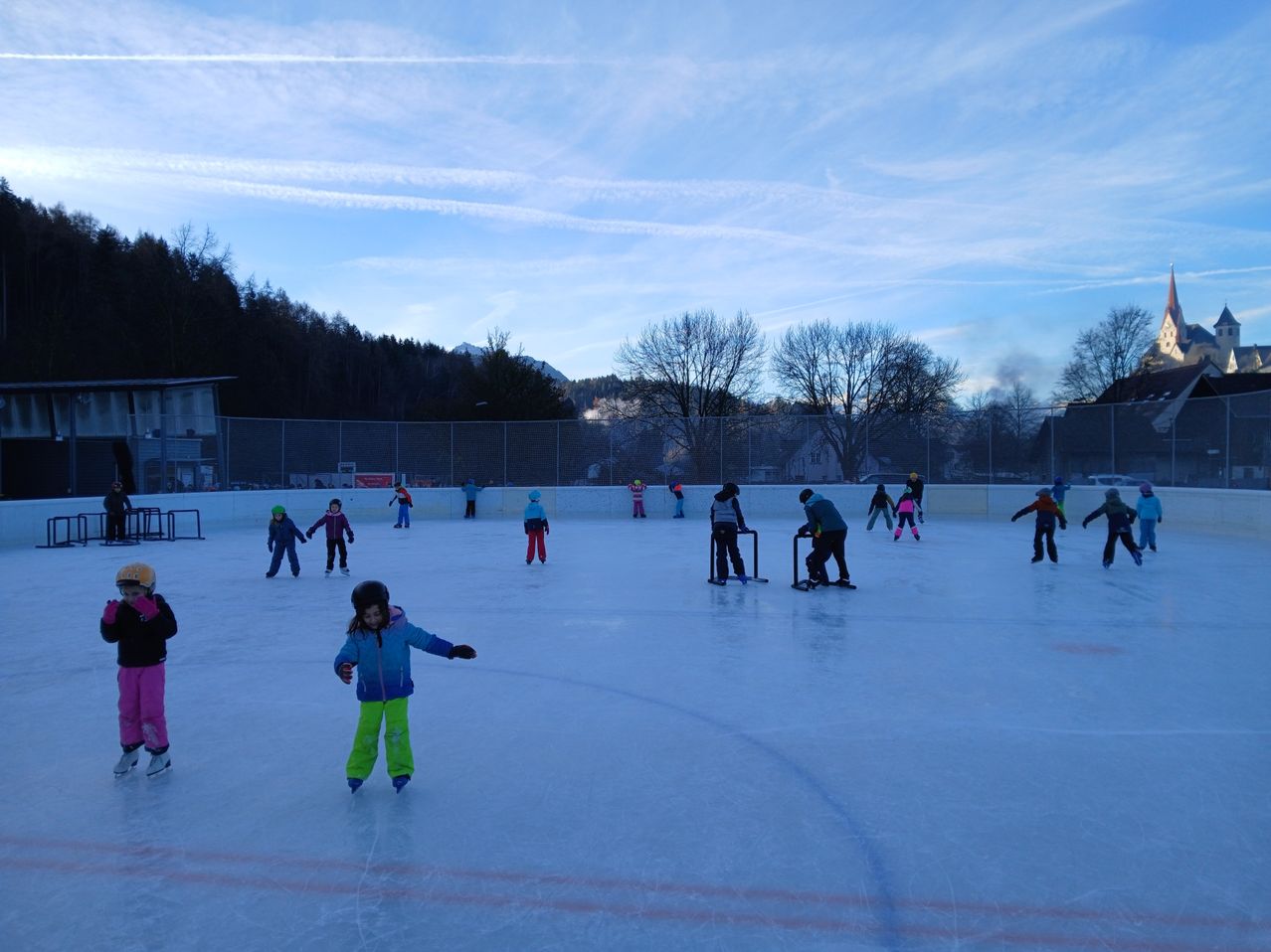 Eine Eislaufbahn im Freien mit mehreren Kindern in Winterkleidung beim Schlittschuhlaufen. Ein Zaun umgibt die Bahn, hinter der Bäume und ein Gebäude sichtbar sind. Der Himmel ist klar mit einigen Wolken.
