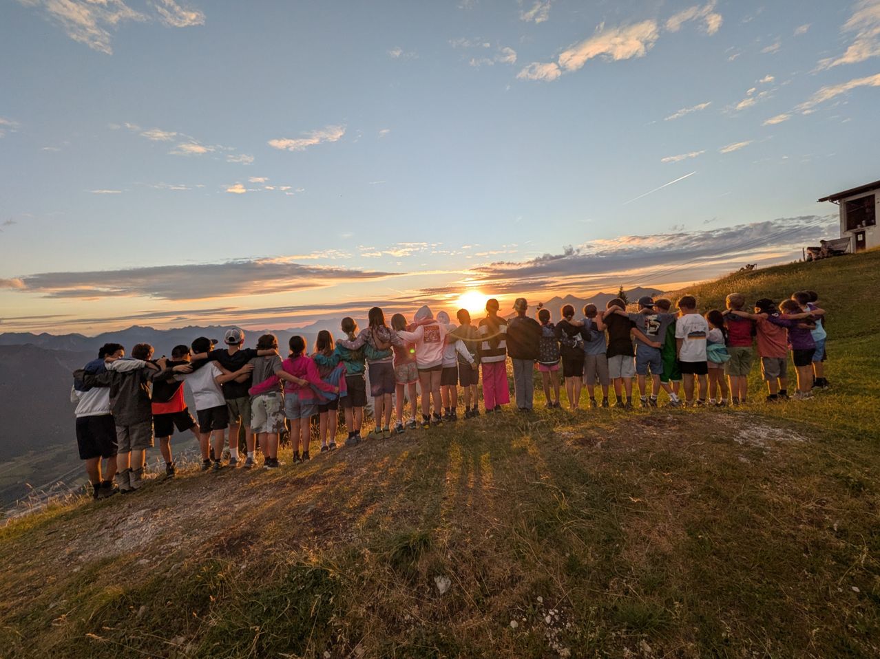 A group of people stand together on a grassy hill, facing the setting sun, with mountains in the background.