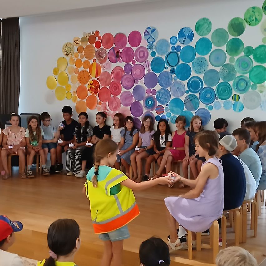 A group of children are seated on stools in front of a colorful wall art, with one child wearing a reflective vest and another holding a small object.