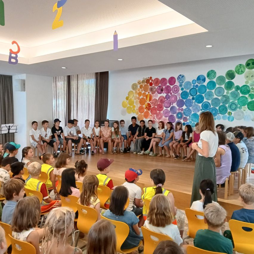 A group of children in yellow vests are sitting in rows of chairs in a classroom, with a woman standing in front of them. The wall behind them is decorated with colorful circles.