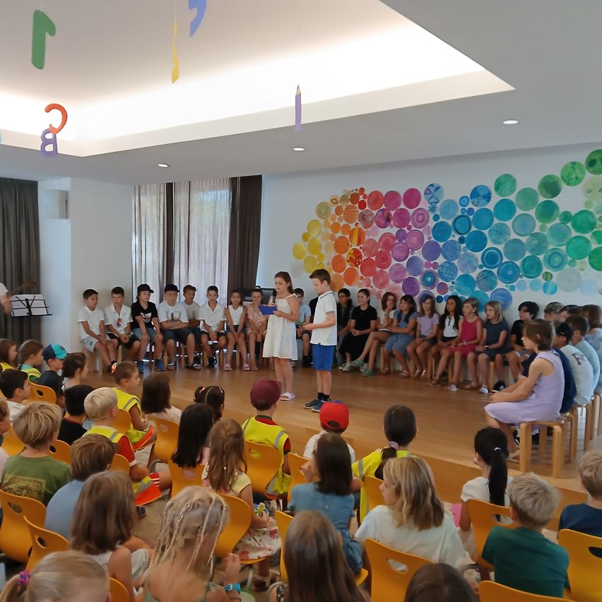 A group of children are sitting in rows on chairs in a classroom with a colorful wall backdrop. A girl stands on stage holding a book, and a boy stands next to her.