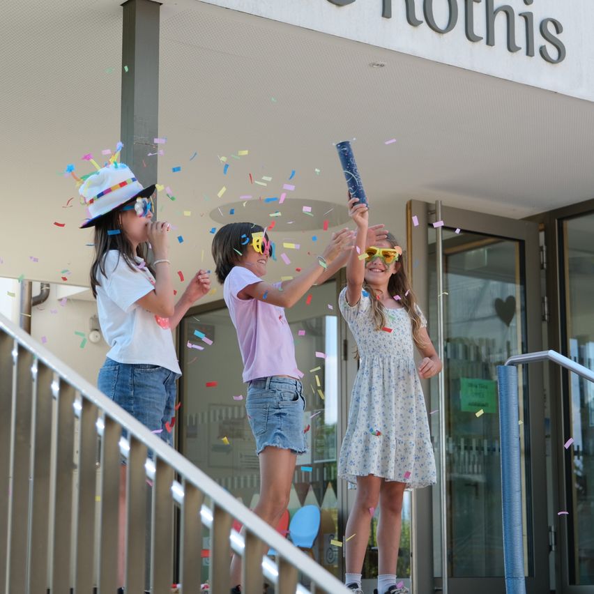 Three girls celebrate with confetti outside a building, one holding a blue tube.