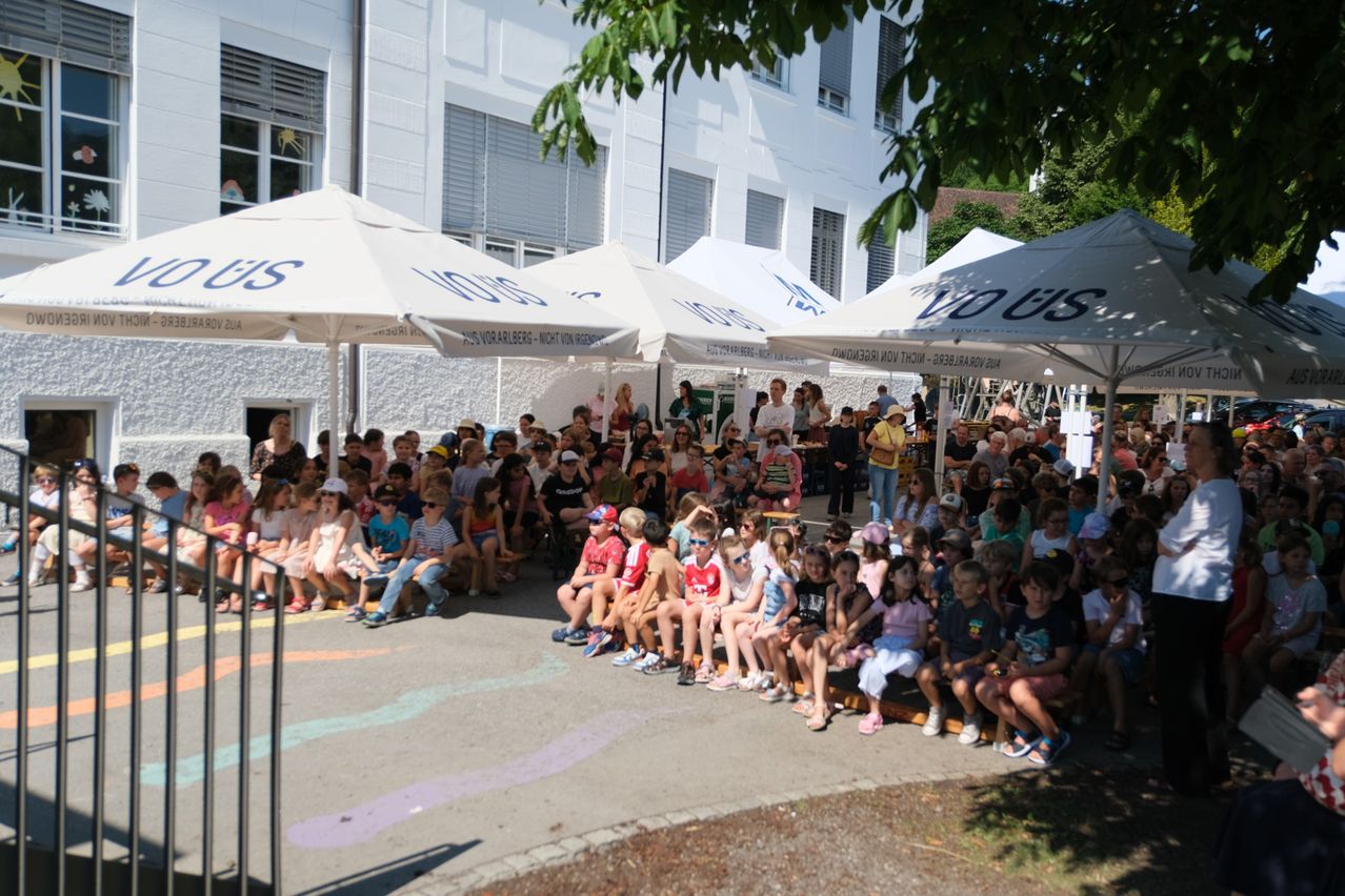A large group of children and adults are seated under white umbrellas in front of a building. Some are wearing hats and glasses. The building has glass windows and is surrounded by trees.