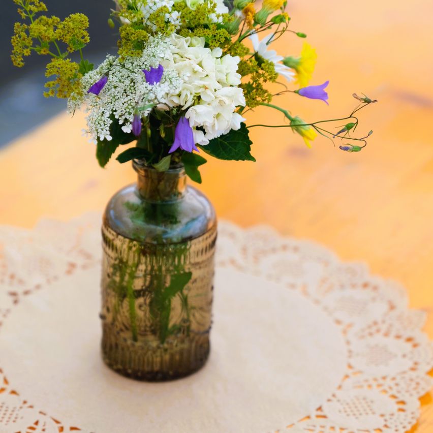 A vintage glass vase with a bouquet of white, yellow, and purple flowers on a doily over a wooden table.