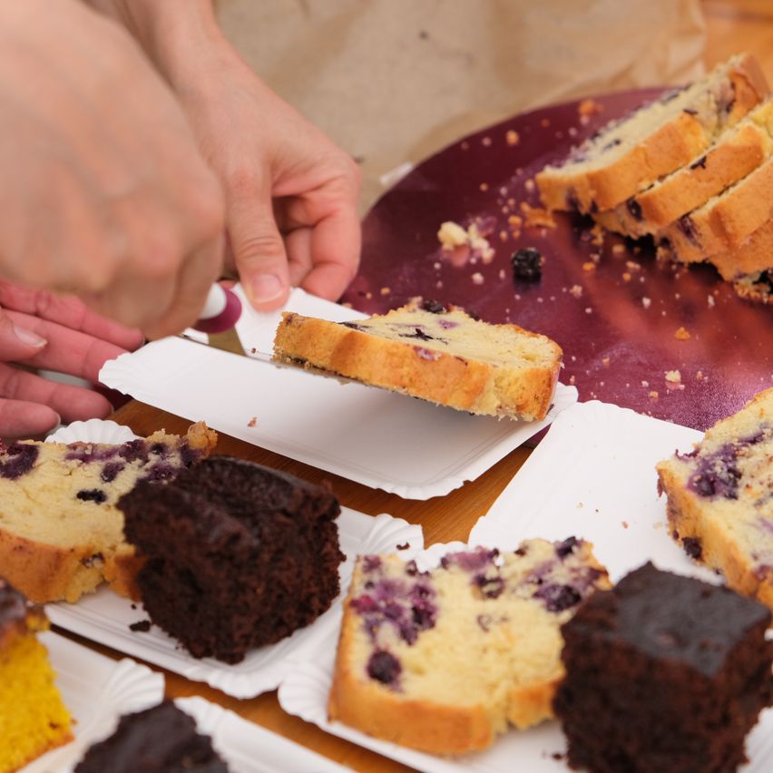 A person slices a piece of blueberry bread from a larger loaf on a table with other desserts.