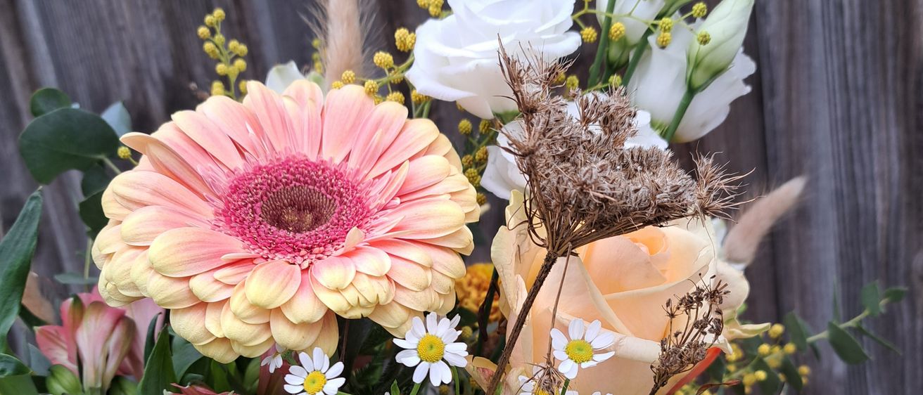 Eine Nahaufnahme eines Blumenstraußes mit verschiedenen Blumen wie Rosen, Gänseblümchen und Gerbera vor einem hölzernen Hintergrund.