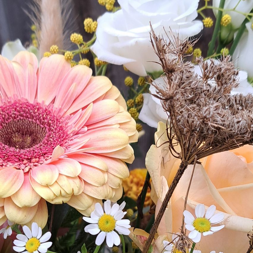 Eine Nahaufnahme eines Blumenstraußes mit verschiedenen Blumen wie Rosen, Gänseblümchen und Gerbera vor einem hölzernen Hintergrund.
