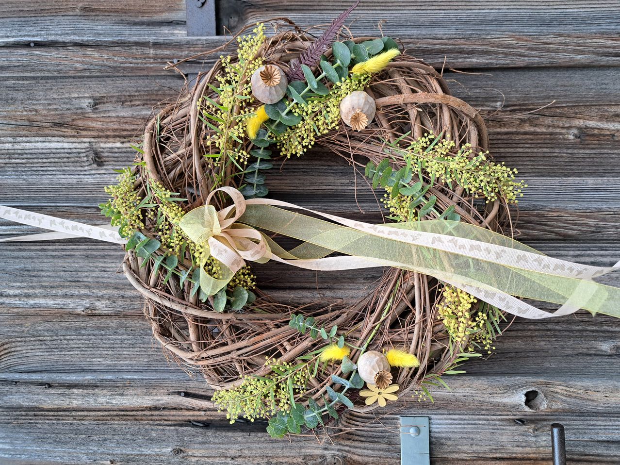A wreath made of brown twigs is adorned with yellow and green flowers, leaves, and a white ribbon. It is displayed on a wooden background.