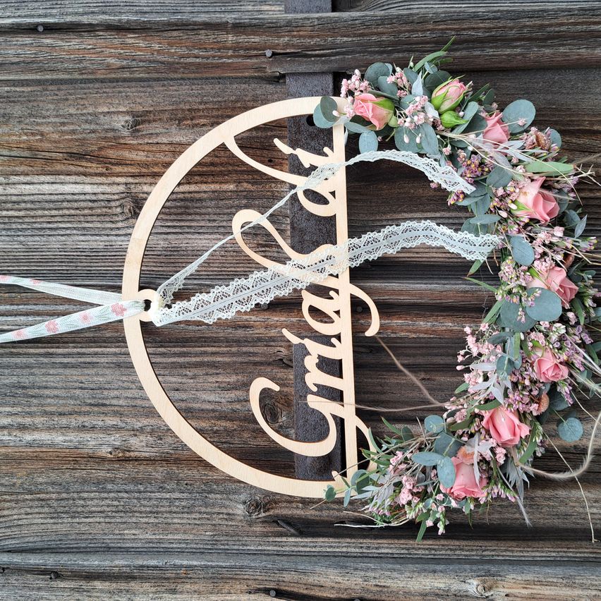A wooden wreath with a name plate 'Amira' and pink flowers and lace is mounted on a wooden wall.