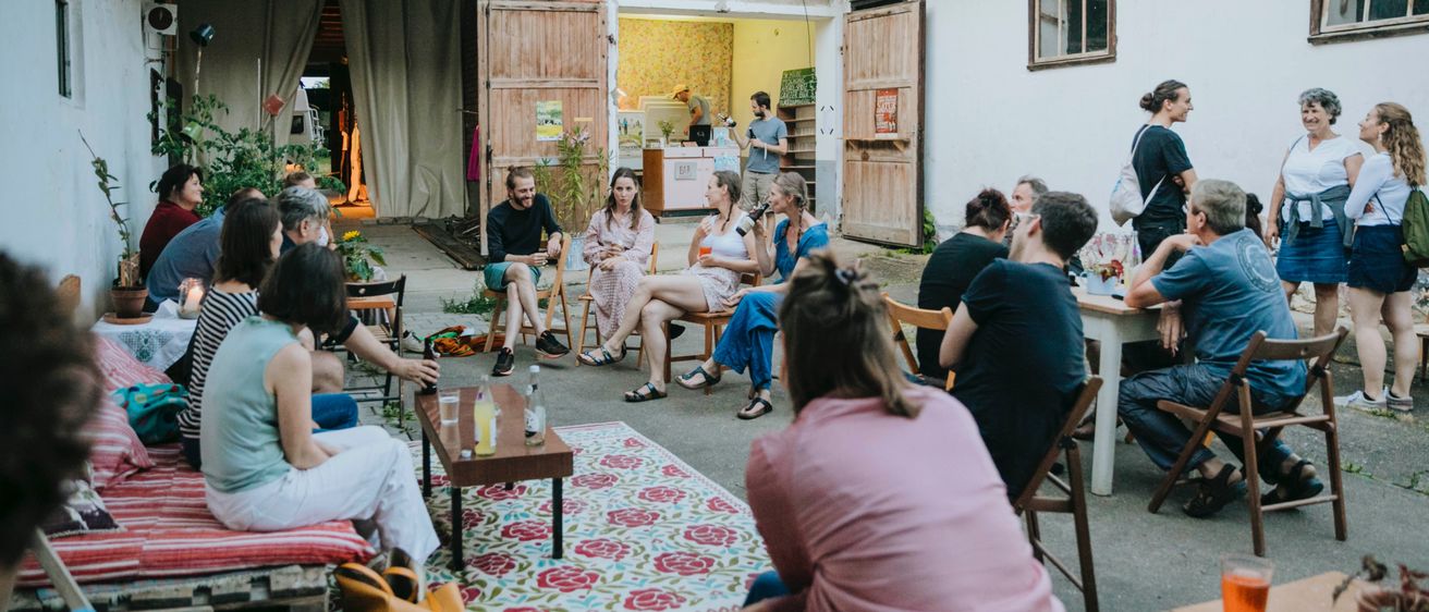 Eine Gruppe von Menschen versammelt sich auf einem Außenhof, sitzt auf Stühlen und Tischen, einige stehen. Sie scheinen in Gespräche vertieft zu sein. Die Umgebung ist rustikal, mit Backsteingebäuden und einem Teppich auf dem Boden.