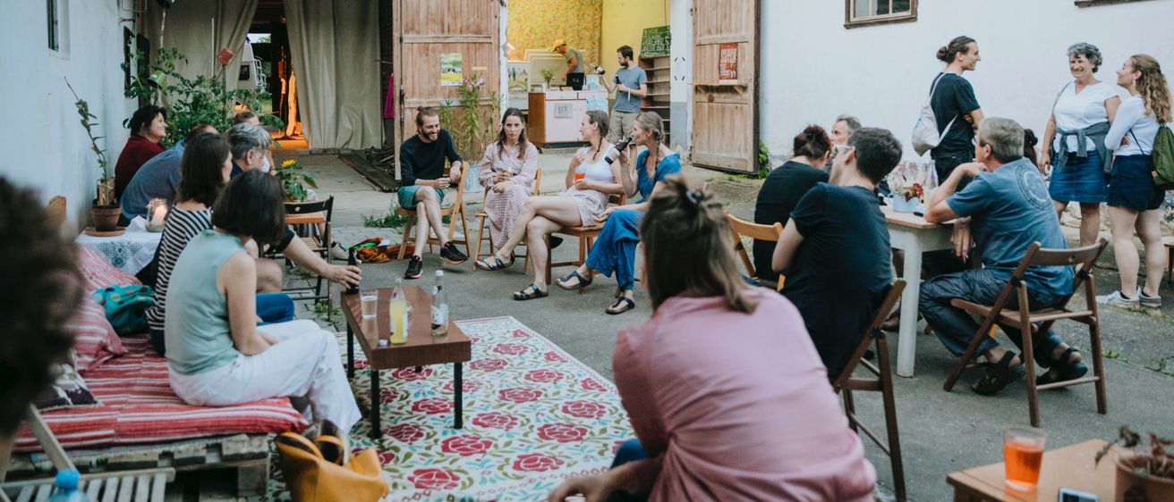 Eine Gruppe von Menschen versammelt sich auf einem Außenhof, sitzt auf Stühlen und Tischen, einige stehen. Sie scheinen in Gespräche vertieft zu sein. Die Umgebung ist rustikal, mit Backsteingebäuden und einem Teppich auf dem Boden.