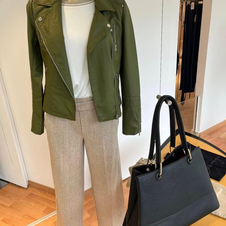 Ein Mannequin präsentiert eine grüne Lederjacke, beige Hosen und eine schwarze Lederhandtasche auf einem Holzboden.
