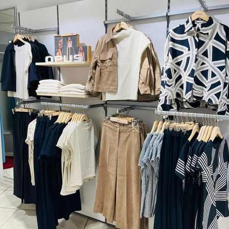 Ein Bekleidungsgeschäft zeigt eine Vielzahl von Outfits an Kleiderbügeln und Regalen, mit verschiedenen Stilen, Farben und Mustern. Im Mittelpunkt steht eine beige Jacke, während die Wände weiß sind und der Boden gefliest ist.