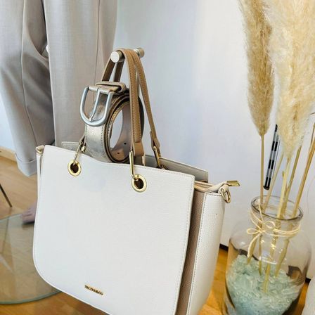 Eine weiße Lederhandtasche mit goldenen Akzenten und einem braunen Riemen steht auf einem Holzboden neben einer Glasvase, die mit Ziersteinen und hohem Pampasgras gefüllt ist.