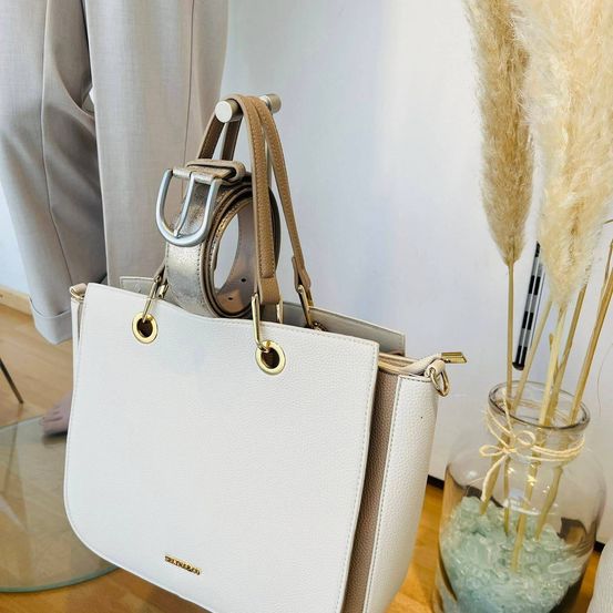 Eine weiße Lederhandtasche mit goldenen Akzenten und einem braunen Riemen steht auf einem Holzboden neben einer Glasvase, die mit Ziersteinen und hohem Pampasgras gefüllt ist.