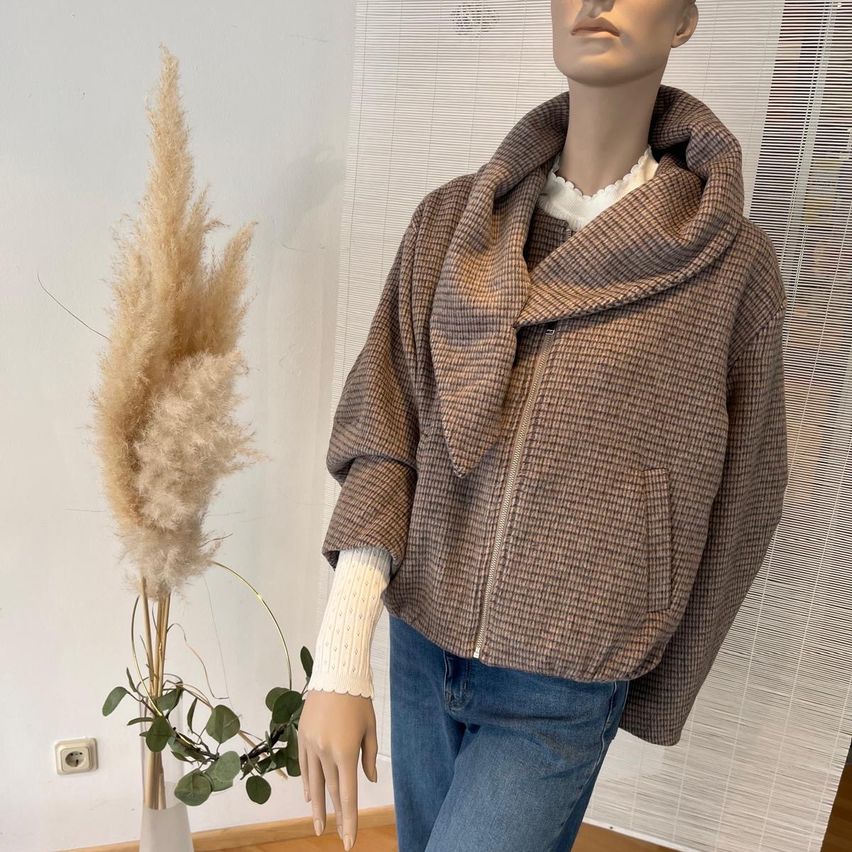 Eine Puppe trägt eine braune karierte Jacke mit Kapuze und blaue Jeans neben einer hohen Pflanze in einem weißen Vasen.