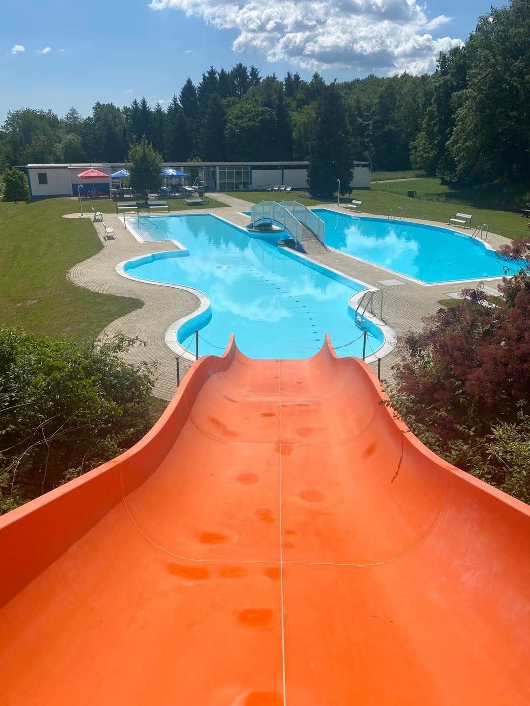 Eine Vogelperspektive auf einen Außenpool mit einem orangefarbenen Wasserrutsche, umgeben von einem Rasenfeld und Gebäuden in der Ferne.