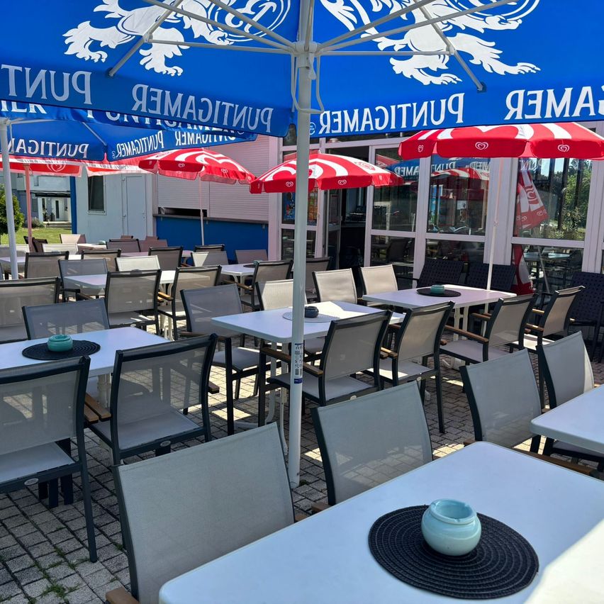 Ein Außenbereich zum Essen mit weißen Tischen und Stühlen. Über jedem Tisch befinden sich Schirme mit weißen und roten Streifen. Ein weißer Topf steht auf einem Tischset vor einem Tisch.