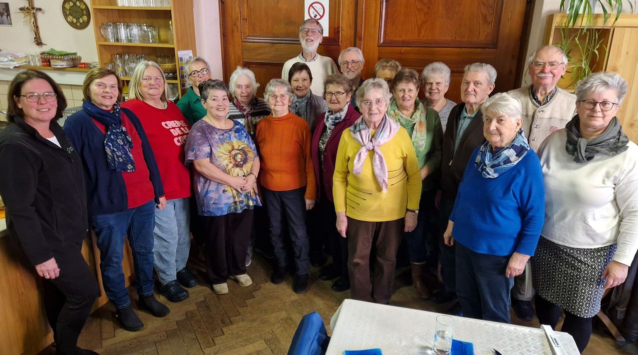 Eine Gruppe älterer Personen, die lächelnd und für ein Foto posieren, in einem Raum mit Holzwänden und einem Tisch.