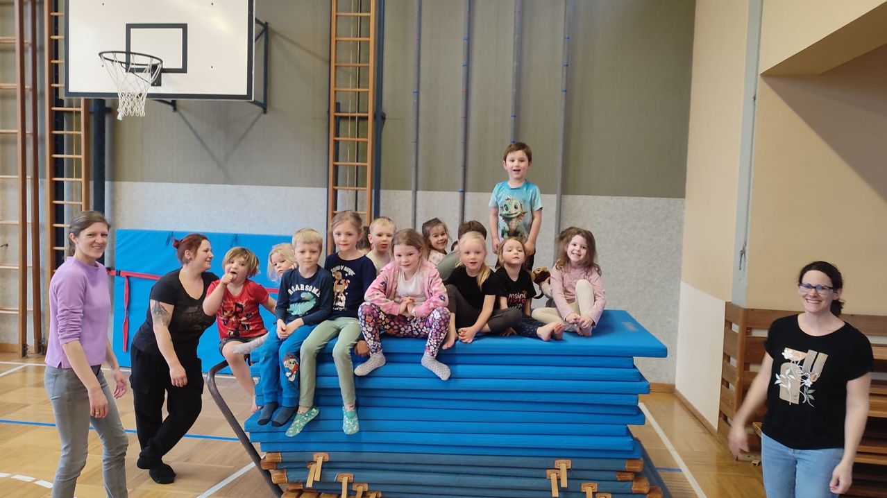 Eine Gruppe von Kindern und einer erwachsenen Frau sitzen auf gestapelten blauen Matten in einer Turnhalle, mit einer Holzleiter im Hintergrund.