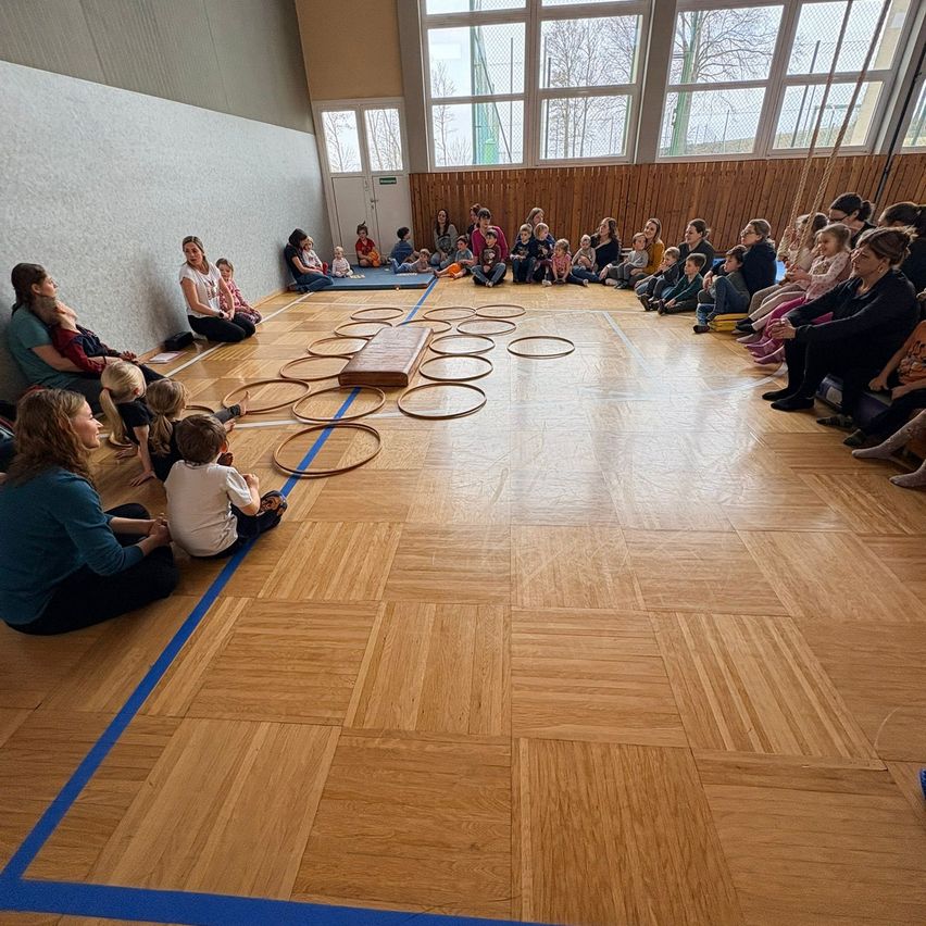 Eine Gruppe von Kindern und Eltern sitzt in einem Kreis um ein Holzringwurfspiel in einer Turnhalle. Blaue Klebeband markiert die Grenzen auf dem Holzboden.