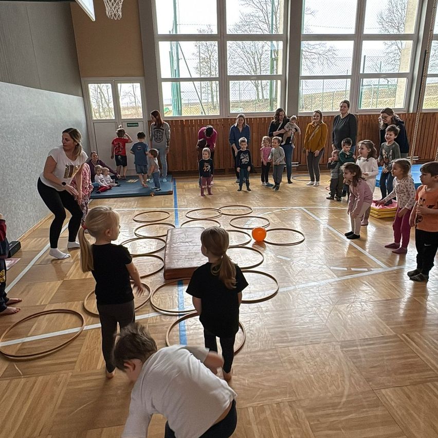 In einer Turnhalle nehmen Kinder an einem Spiel mit Hula-Hoops auf einem Holzboden teil. Eine Frau leitet die Aktivität, während Eltern und andere Erwachsene von den Seitenlinien aus zusehen.