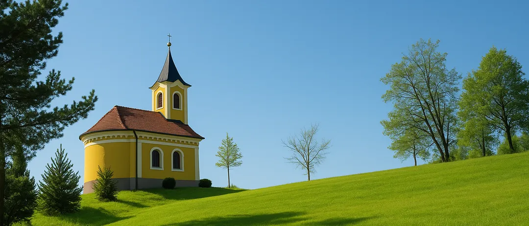 Eine kleine gelbe Kapelle steht auf einem grünen Hügel unter einem klaren blauen Himmel mit ein paar Bäumen in der Nähe.