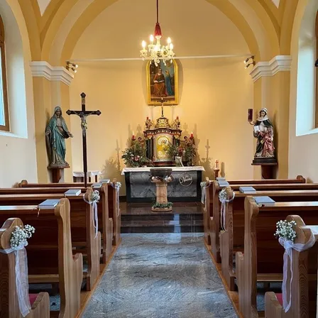 Ein Kircheninneres zeigt eine Kronleuchter, ein Kreuz und Statuen auf beiden Seiten des Altars. Die Kirchenbänke sind mit weißen Blumen geschmückt, und ein Teppich führt zum Altar.