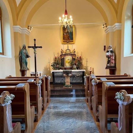 Ein Kircheninneres zeigt eine Kronleuchter, ein Kreuz und Statuen auf beiden Seiten des Altars. Die Kirchenbänke sind mit weißen Blumen geschmückt, und ein Teppich führt zum Altar.