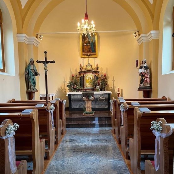 Ein Kircheninneres zeigt eine Kronleuchter, ein Kreuz und Statuen auf beiden Seiten des Altars. Die Kirchenbänke sind mit weißen Blumen geschmückt, und ein Teppich führt zum Altar.