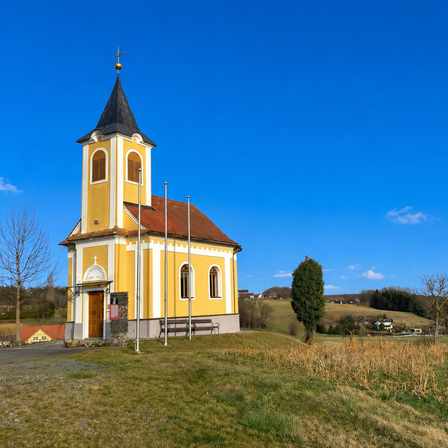 Eine kleine gelbe Kirche mit einem schwarzen Turm steht auf einem Hügel mit ein paar Bäumen und einer Bank davor. Der Himmel ist klar blau.