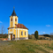 Eine kleine gelbe Kirche mit einem schwarzen Turm steht auf einem Hügel mit ein paar Bäumen und einer Bank davor. Der Himmel ist klar blau.