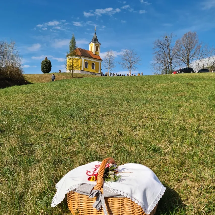 Ein Korb mit einem weißen Tuch und Blumen steht auf einem Hügel mit einer gelben Kirche im Hintergrund unter einem blauen Himmel.