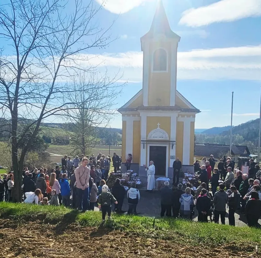 Ein Priester spricht zu einer Menschenmenge vor einer gelben Kirche unter einem teilweise bewölkten Himmel. Eine große Gruppe von Menschen versammelt sich in der Nähe von Tischen, die vor der Kirche aufgestellt sind.