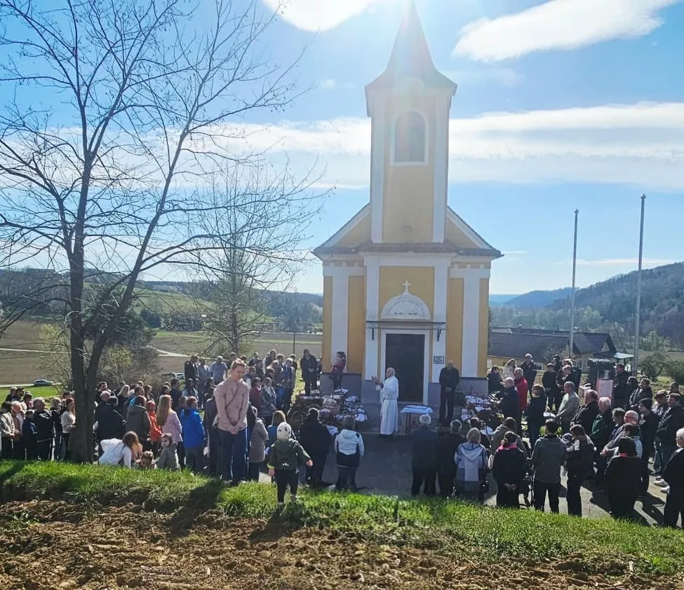 Ein Priester spricht zu einer Menschenmenge vor einer gelben Kirche unter einem teilweise bewölkten Himmel. Eine große Gruppe von Menschen versammelt sich in der Nähe von Tischen, die vor der Kirche aufgestellt sind.