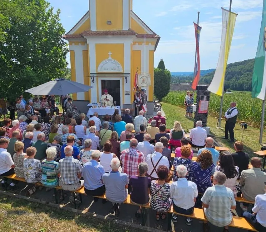Eine Gruppe von Menschen versammelt sich vor einer kleinen Kapelle für eine religiöse Zeremonie. Der Priester steht am Altar, und mehrere Flaggen sind an Stangen angebracht. Die Teilnehmer sitzen auf Bänken, und einige stehen. Die Kapelle ist von Grün umgeben und in der Ferne sind Berge zu sehen.