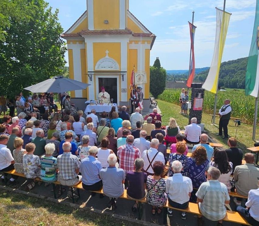 Eine Gruppe von Menschen versammelt sich vor einer kleinen Kapelle für eine religiöse Zeremonie. Der Priester steht am Altar, und mehrere Flaggen sind an Stangen angebracht. Die Teilnehmer sitzen auf Bänken, und einige stehen. Die Kapelle ist von Grün umgeben und in der Ferne sind Berge zu sehen.