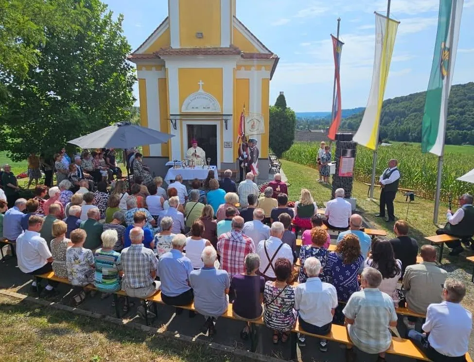 Eine Gruppe von Menschen versammelt sich vor einer kleinen Kapelle für eine religiöse Zeremonie. Der Priester steht am Altar, und mehrere Flaggen sind an Stangen angebracht. Die Teilnehmer sitzen auf Bänken, und einige stehen. Die Kapelle ist von Grün umgeben und in der Ferne sind Berge zu sehen.
