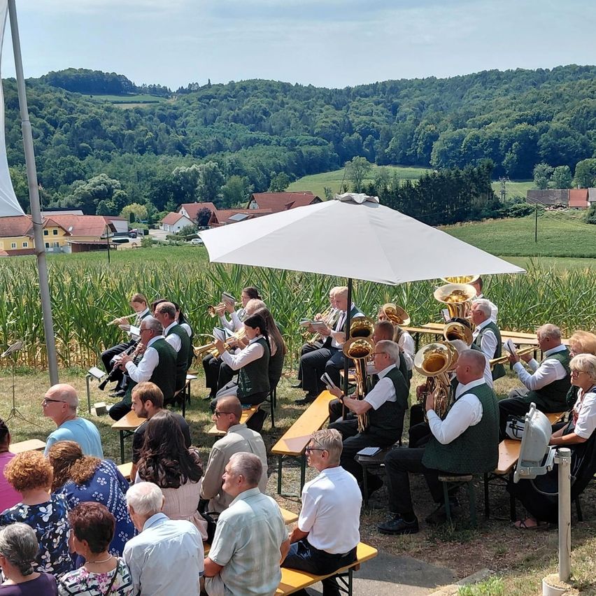 Eine Blaskapelle spielt im Freien auf Bänken unter einem weißen Schirm, mit einer malerischen Kulisse aus Maisfeld und Hügeln.