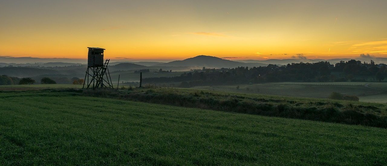 Ein hölzerner Aussichtsturm steht in einem üppigen grünen Feld, während die Sonne untergeht und einen warmen Schimmer über die nebligen Berge in der Ferne wirft.