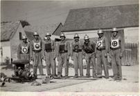 Ein Schwarz-Weiß-Foto von sieben Männern in Uniform und Helmen, die in einer Reihe stehen, wahrscheinlich in einer militärischen Umgebung.