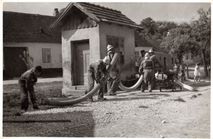 Schwarz-Weiß-Foto von Feuerwehrleuten, die einen Schlauch benutzen, um ein Feuer vor einem Haus zu löschen.
