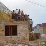 Eine Gruppe von Arbeitern, alle mit Hüten, steht auf einem im Bau befindlichen Backsteingebäude. Sie stehen auf dem Dach und halten Holzbretter. Ein Mann steht auf einer Leiter.